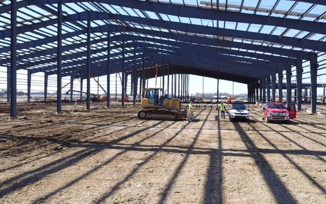 Steel-frame building under construction with vehicles, workers, cars.