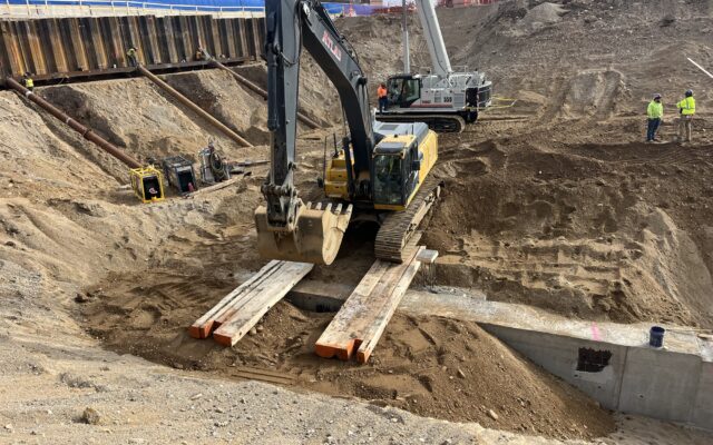 Excavator crosses planks over trench as workers watch.