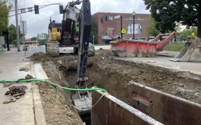 Excavator digging trench by city street with barriers and buildings.