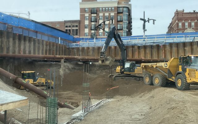 Excavators and dump trucks work in a deep urban construction pit.
