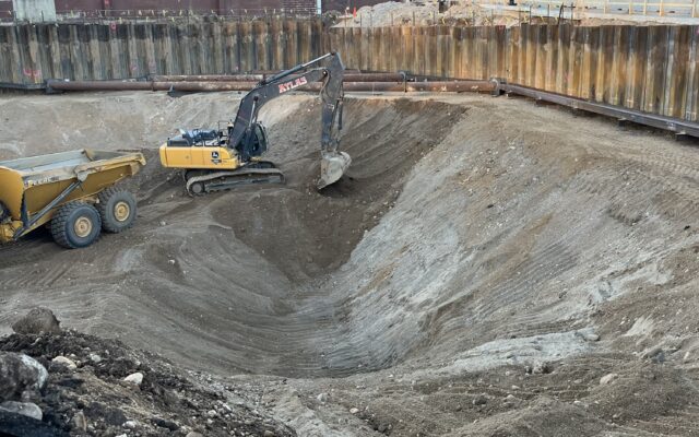 Excavator and dump truck in pit, city buildings behind.