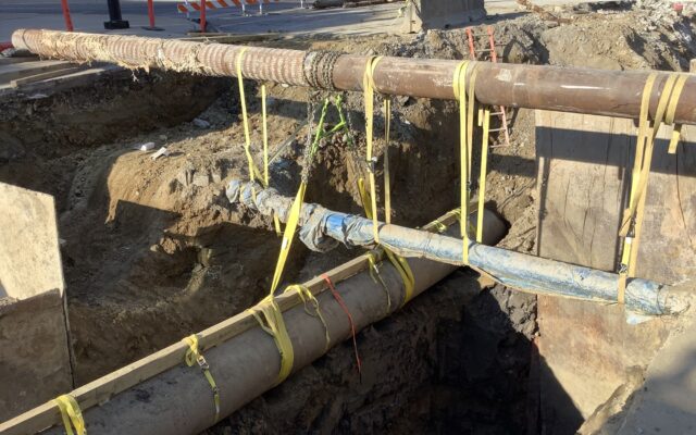 Three large pipes with yellow straps over a trench; barriers visible.