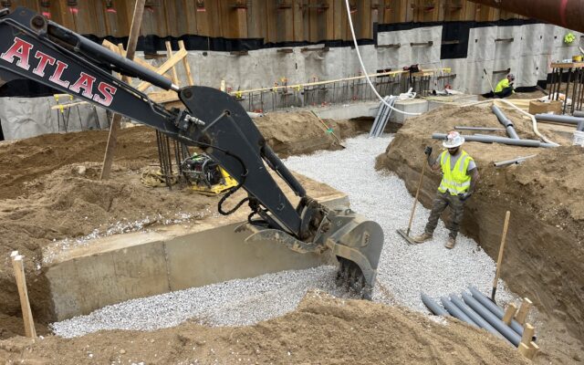 Worker in gravel trench near excavator, pipes and materials visible.