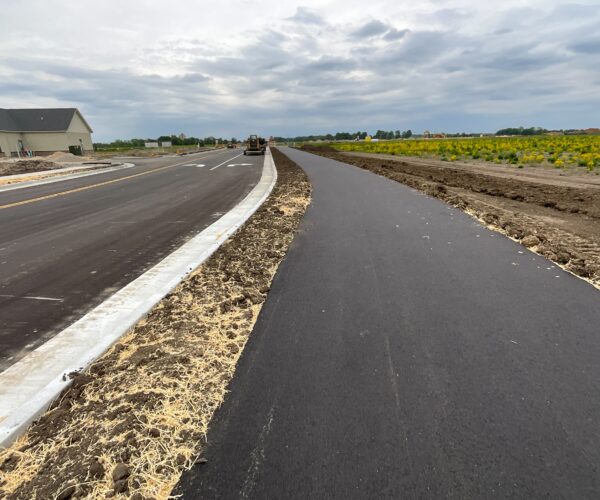 New asphalt bike path beside road, bare soil, cloudy sky.
