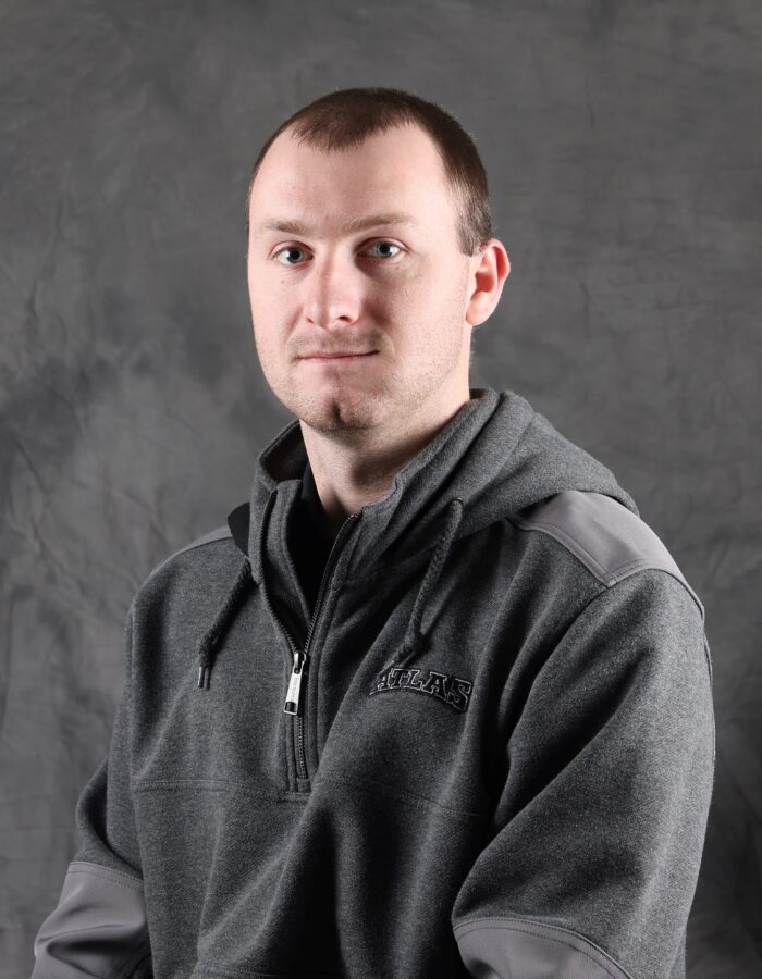 Man in gray hoodie sits before gray backdrop, neutral expression.