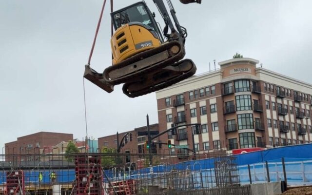 Crane lifts small excavator as workers watch on construction site.
