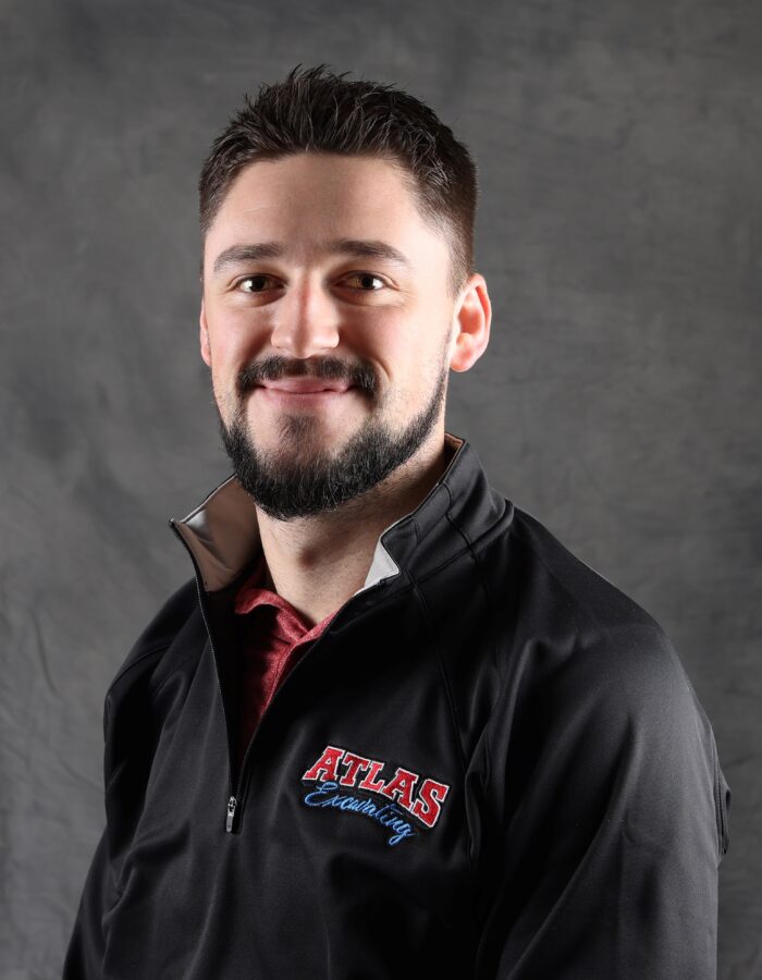 Smiling man with beard and "Atlas" jacket before gray backdrop.