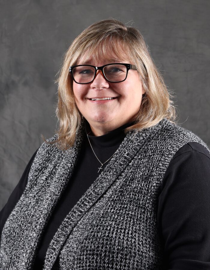 Blonde woman with glasses, black top, gray vest on gray background.
