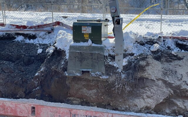 Green utility box and damaged meter on snowy site behind barrier.