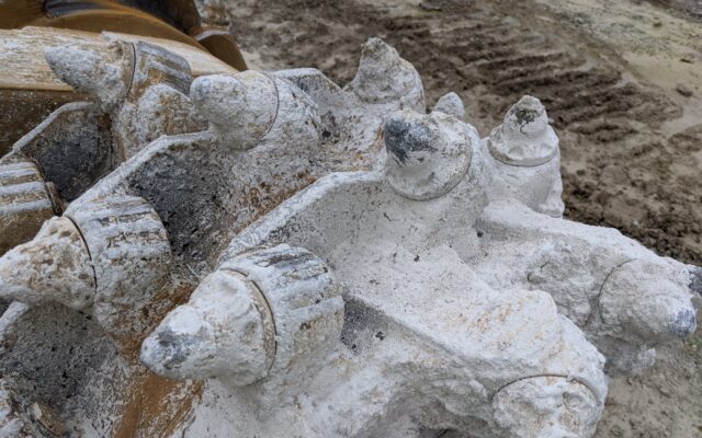 Dusty rotary excavator drum with worn teeth at construction site.
