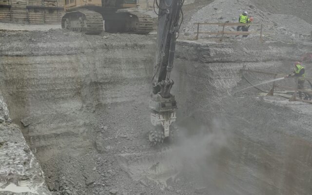 Excavator moves rocks in pit as two workers watch nearby.