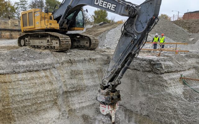 Excavator with grinder works in pit; workers watch.