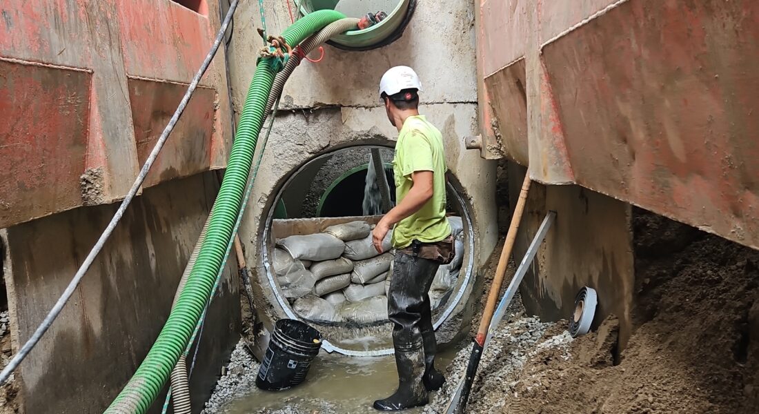 Worker in muddy trench with pipes, sandbags, hoses; red walls.
