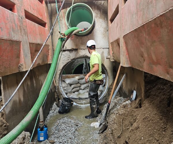 Worker in muddy trench with pipes, sandbags, hoses; red walls.