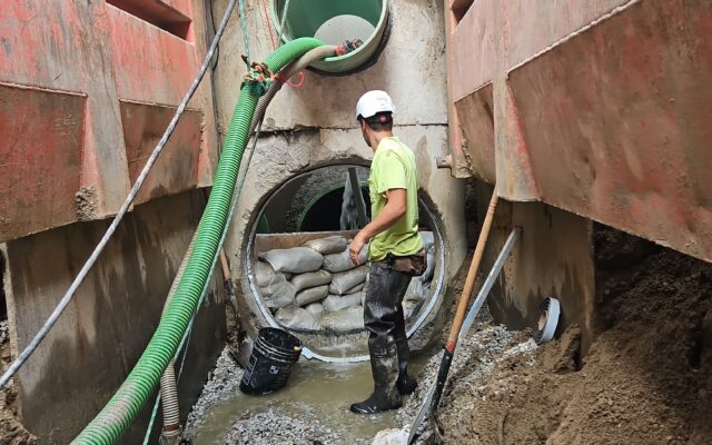 Worker in muddy trench with pipes, sandbags, hoses; red walls.
