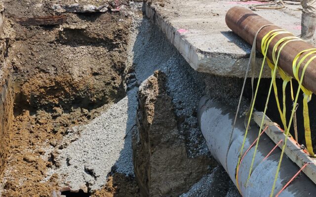 Excavator and worker beside trench with exposed pipes, timestamp.