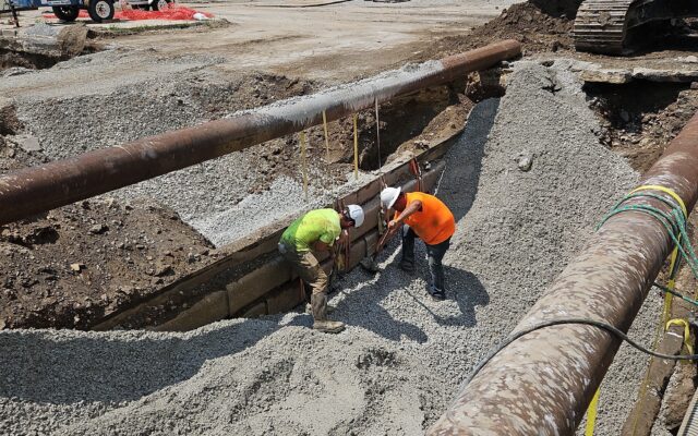 Two workers in safety gear near large pipes at construction site.