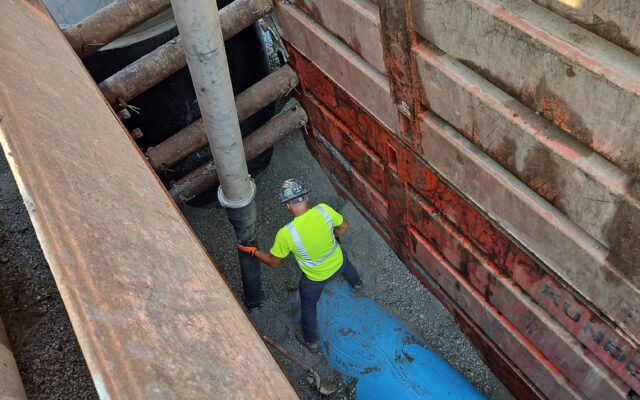 Worker in safety gear by blue pipe in deep, steel-walled site.