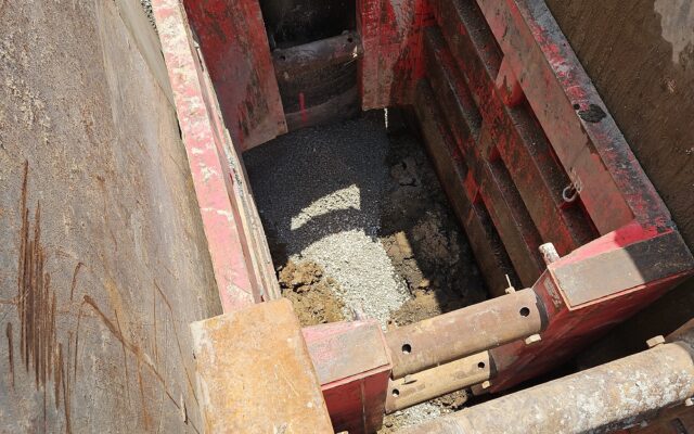 Red trench box by gravel, concrete manhole; 7/15/24 timestamp.
