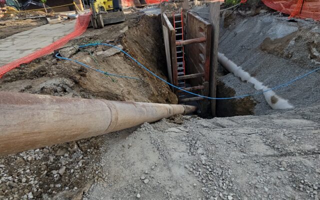 Excavated trench, machinery, large pipes; fenced construction site. Timestamp: 7/29/24, 8:07:35 AM EDT.