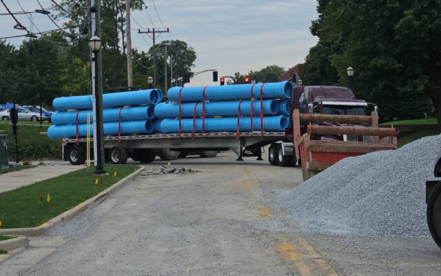 Flatbed truck with blue pipes parked on gravel street under construction.