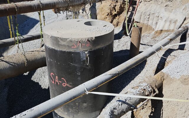Construction site with concrete cylinder, pipes, gravel, and trench.