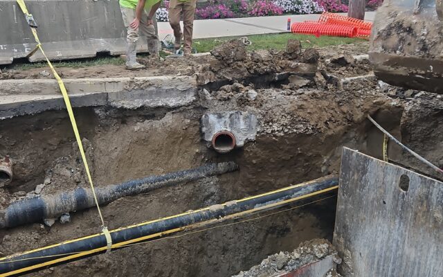 Workers by open trench and pipes, site cordoned off, morning.