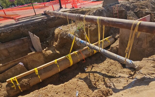 Excavated site with pipes, yellow straps, orange fencing, barriers.