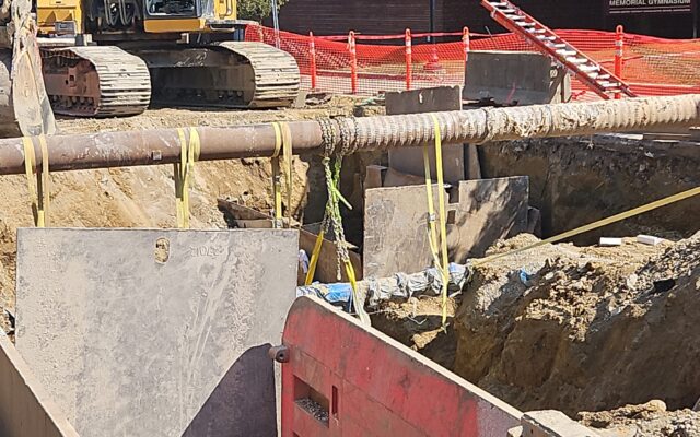 Excavator, trench shields, pipes, and safety fencing by brick building.