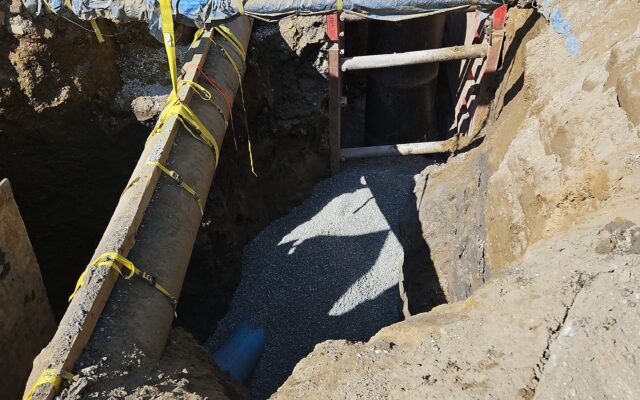 Construction trench with large pipes, yellow straps, gravel, metal supports.