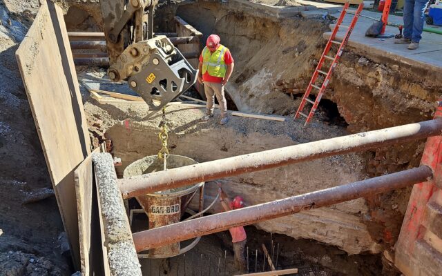 Construction workers use heavy machinery in a city street excavation.