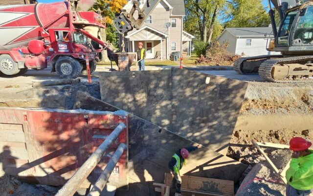 Workers install pipe underground; excavator, cement mixer, date/time shown.