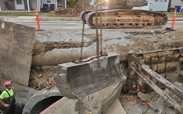 Excavator lifts slab by large pipe; worker in safety gear nearby.