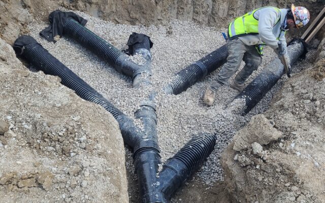 Worker in safety gear installs drainage pipes in gravel trench.