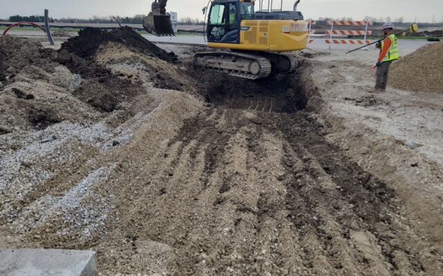 Excavator digs near worker; dirt piles and barriers visible.