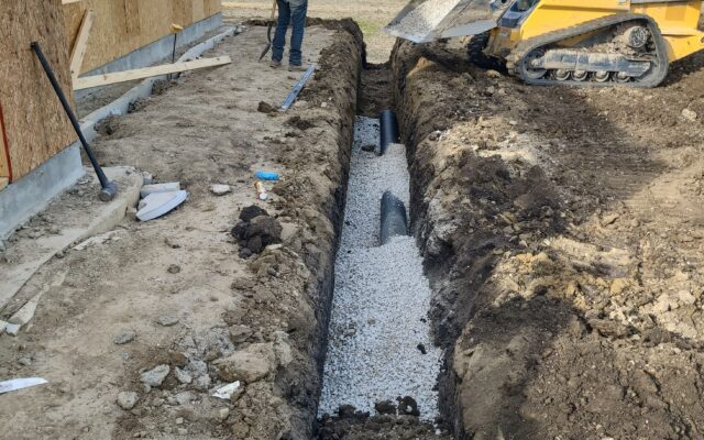 Worker guides loader near trench, pipes, gravel, and building frame.