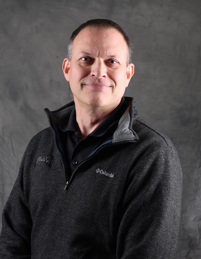 Man with short hair in dark fleece, smiling against gray background.