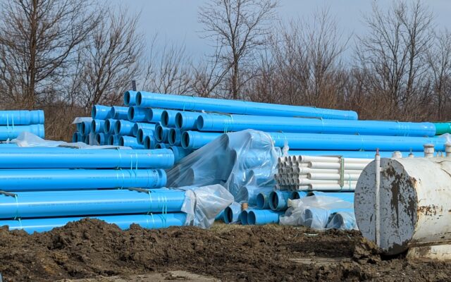 Plastic pipes stacked by a white tank, bare trees behind.