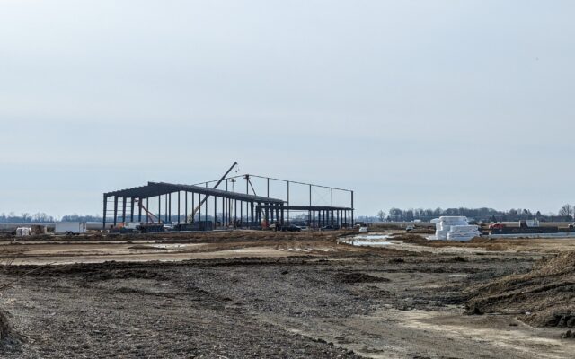 Steel frame rising on dirt lot, crane and materials nearby.