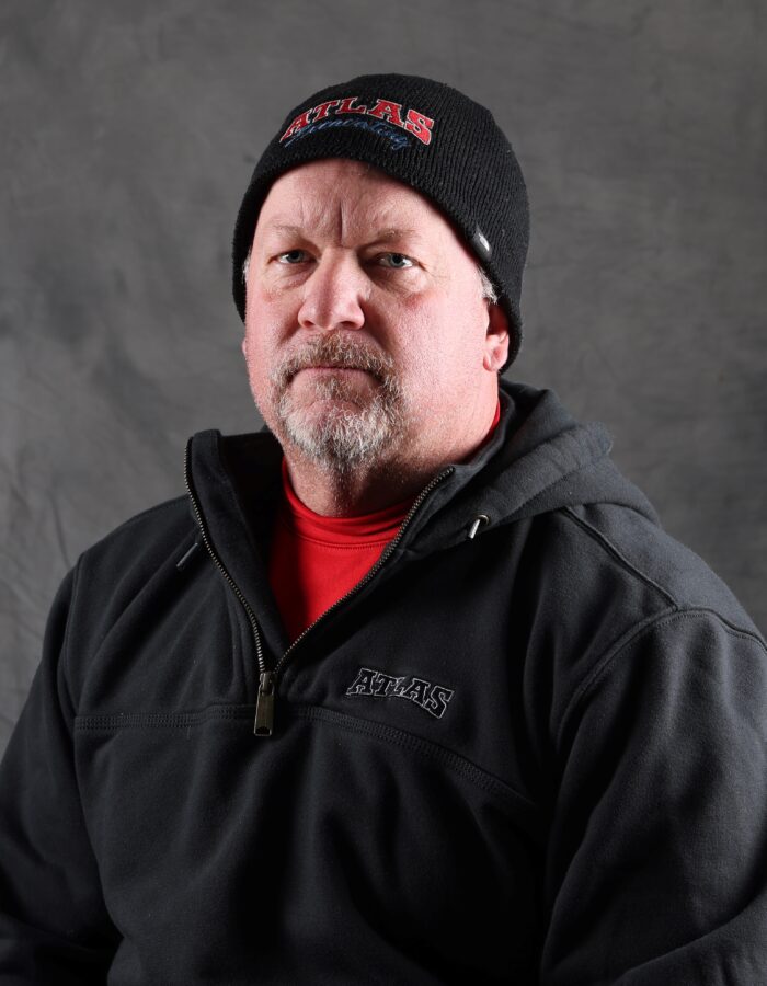 Man with short beard in black beanie and jacket, gray background.