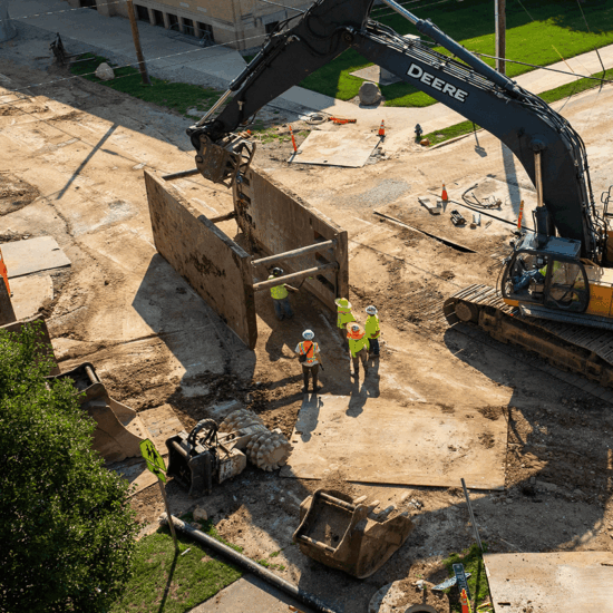 Workers and excavator move trench box on dirt road; cones nearby.