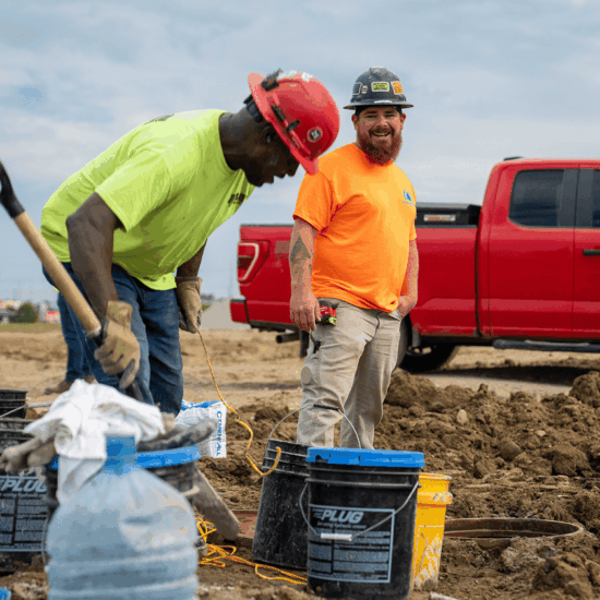 Two construction workers at site; one digs, one smiles; red truck behind.