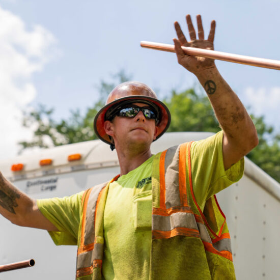 Worker in hard hat and vest lifts copper pipe outdoors.