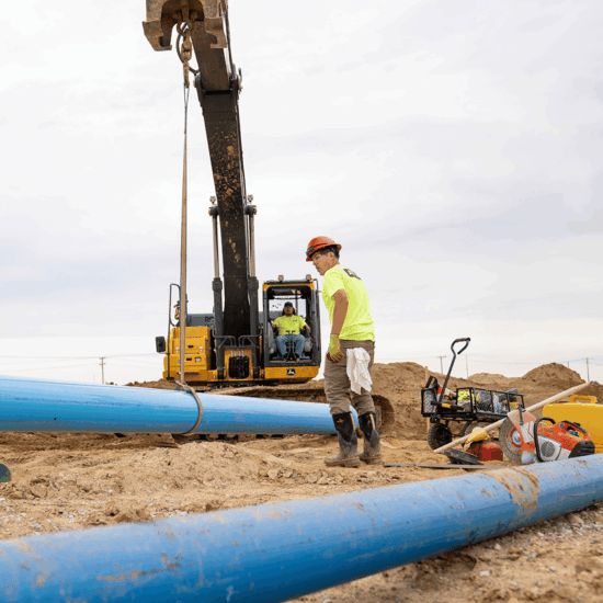 Worker in hard hat watches excavator lift blue pipe.