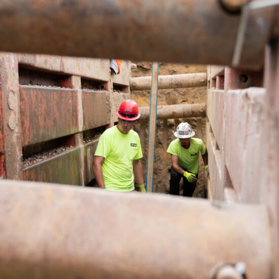 Two workers in safety gear stand inside a reinforced trench.