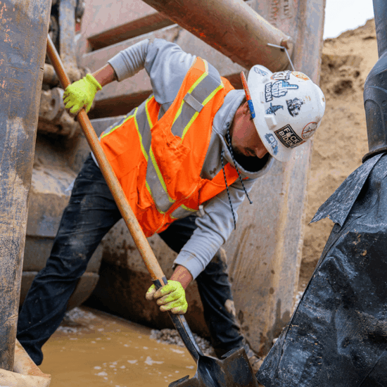 Worker in orange vest digs with shovel near machinery.