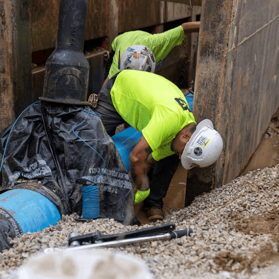 Workers in safety gear inspect large blue pipes in trench.